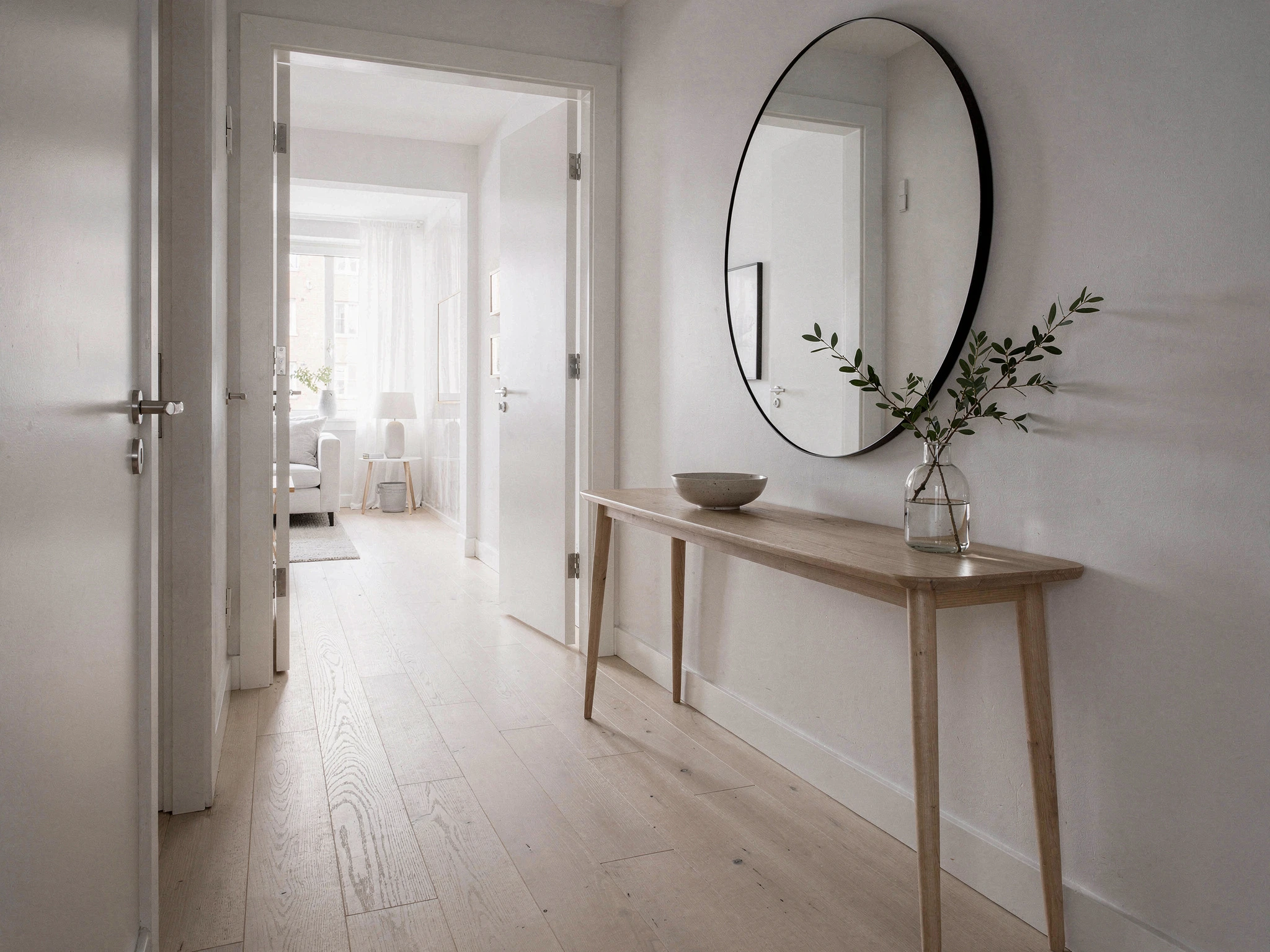 Hall med ljust trägolv, avlastningsbord och rund spegel